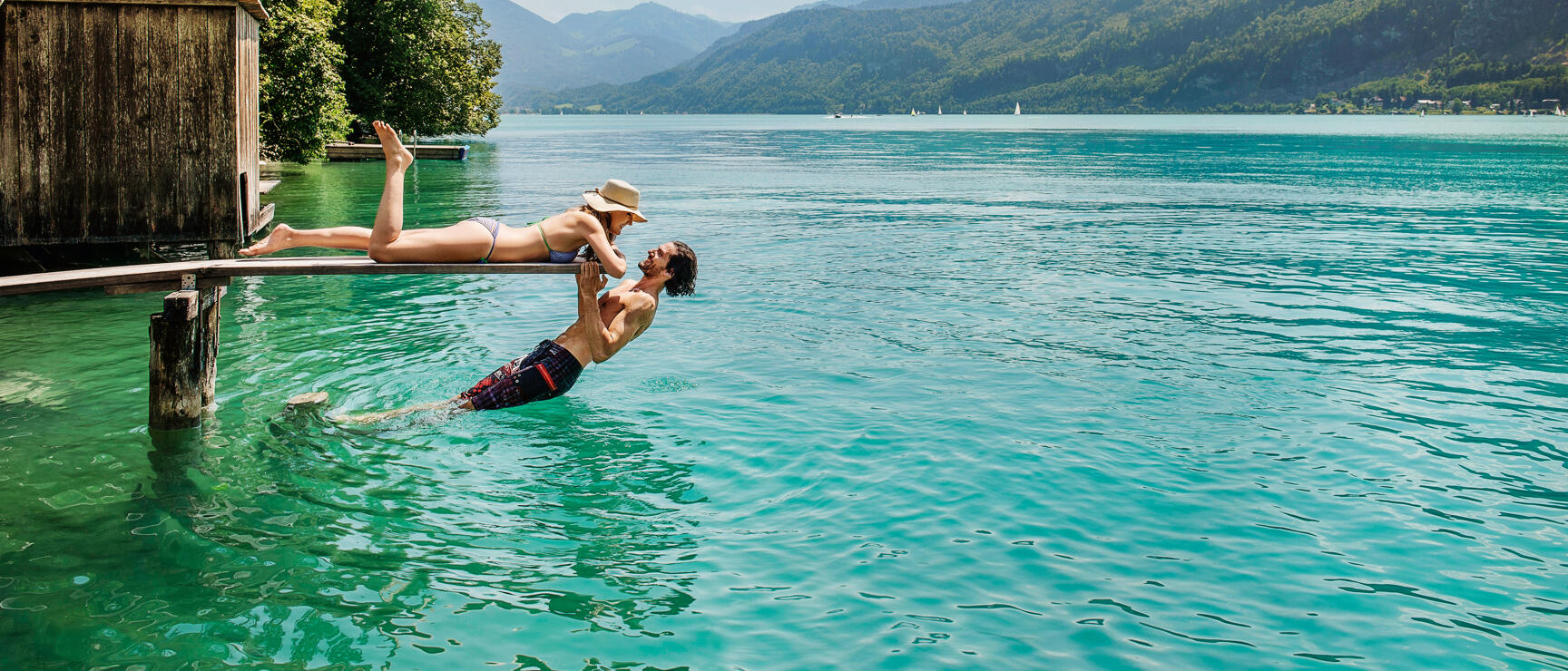 Woman in straw hat lying on jetty, man in water holding her, turquoise lake with mountains in background.