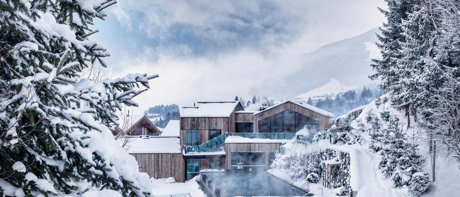 Wooden hotel with steaming outdoor pool in snow, surrounded by snowy fir trees and mountains in the background.