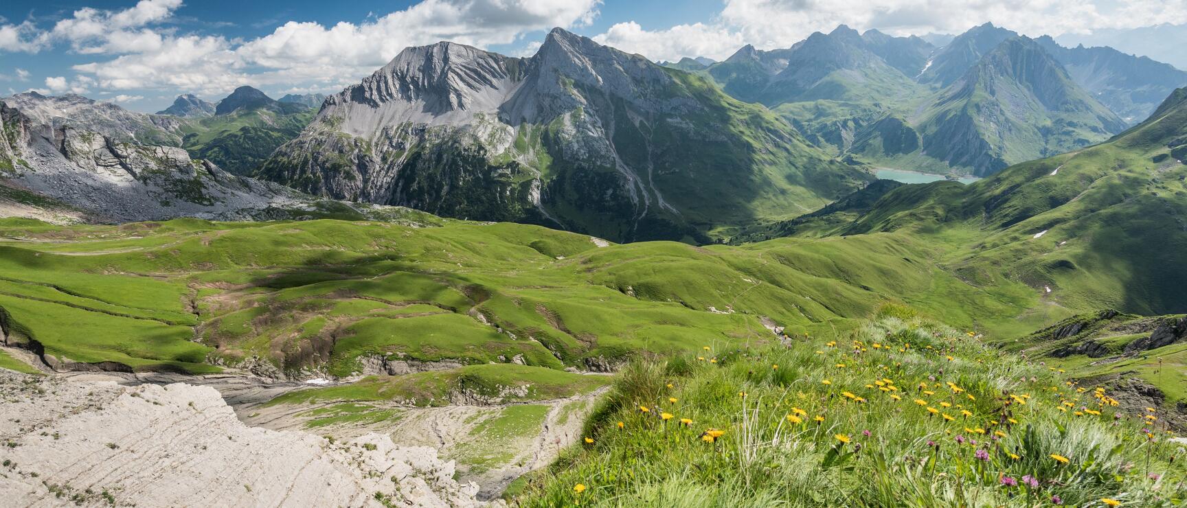 Wide mountain panorama with green alpine meadows, yellow flowers, rock formations and lake.