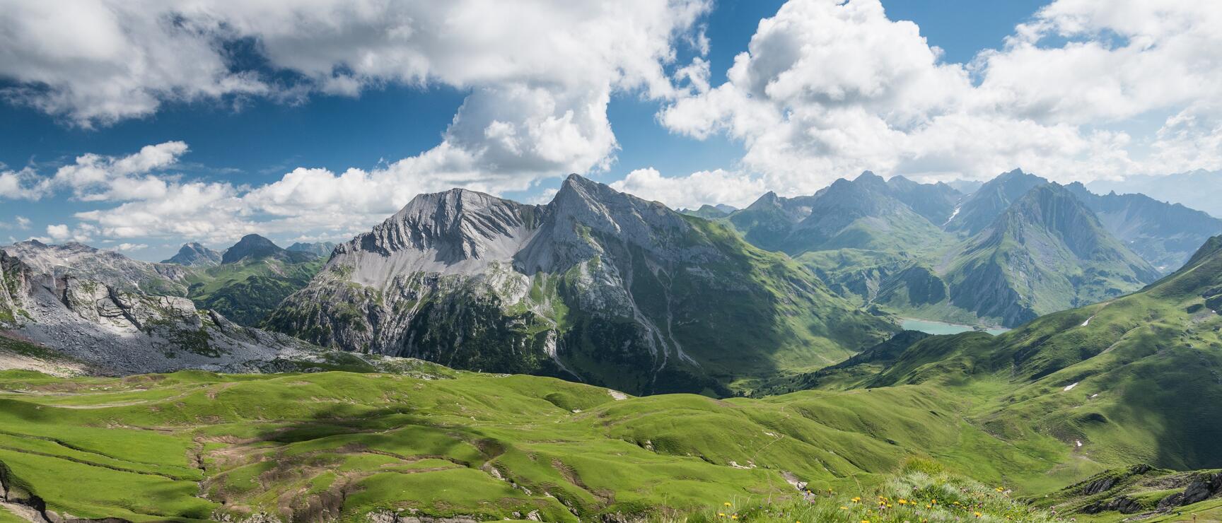 Weites Bergpanorama mit grünen Almwiesen, gelben Blumen, Felsformationen und einem See im Hintergrund.