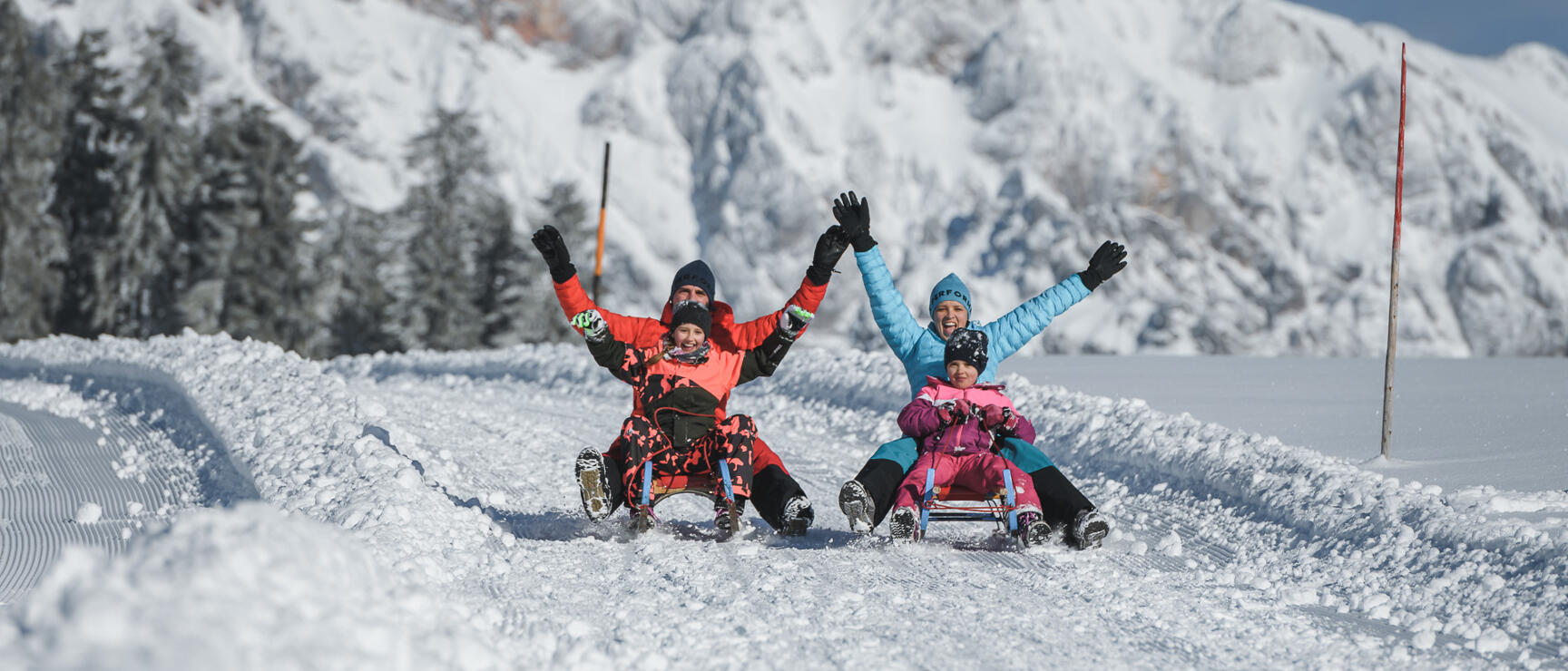 Four people on sledges on a toboggan run with hands raised in the air, snow-covered mountains in background.