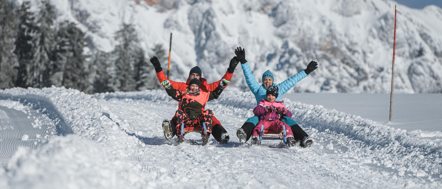 Four people on sledges on a toboggan run with hands raised in the air, snow-covered mountains in background.