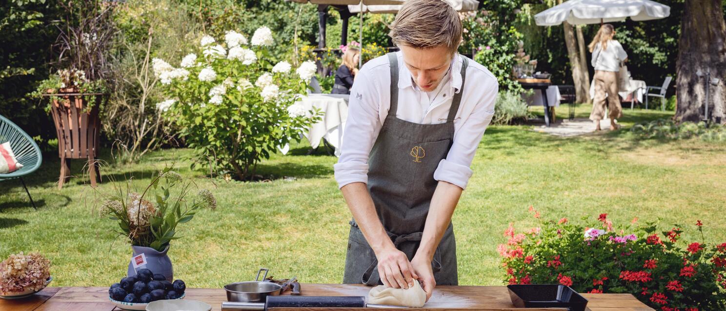 Chef in grey apron kneading dough at a wooden table in the garden with plums and baking pans nearby.