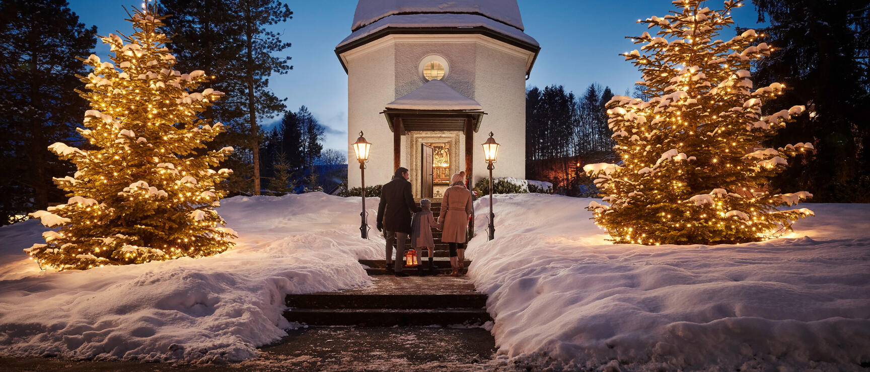 Silent Night Chapel in Oberndorf in winter, two adults and child at entrance with illuminated Christmas trees.