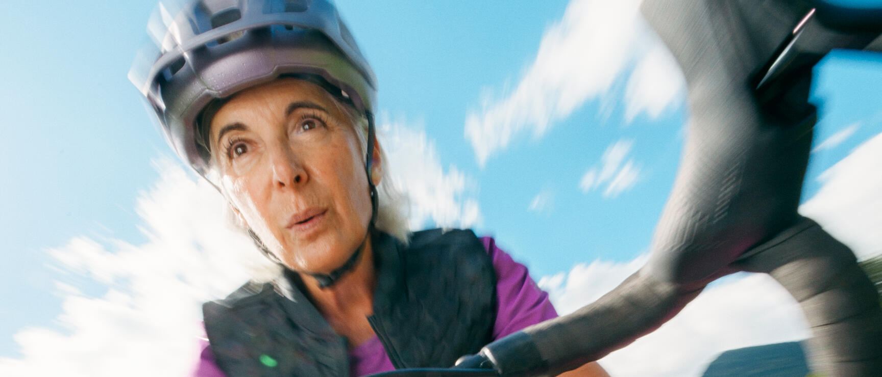 Woman wearing a helmet on a bicycle, close-up of the handlebars, green meadows and blue sky in the background.