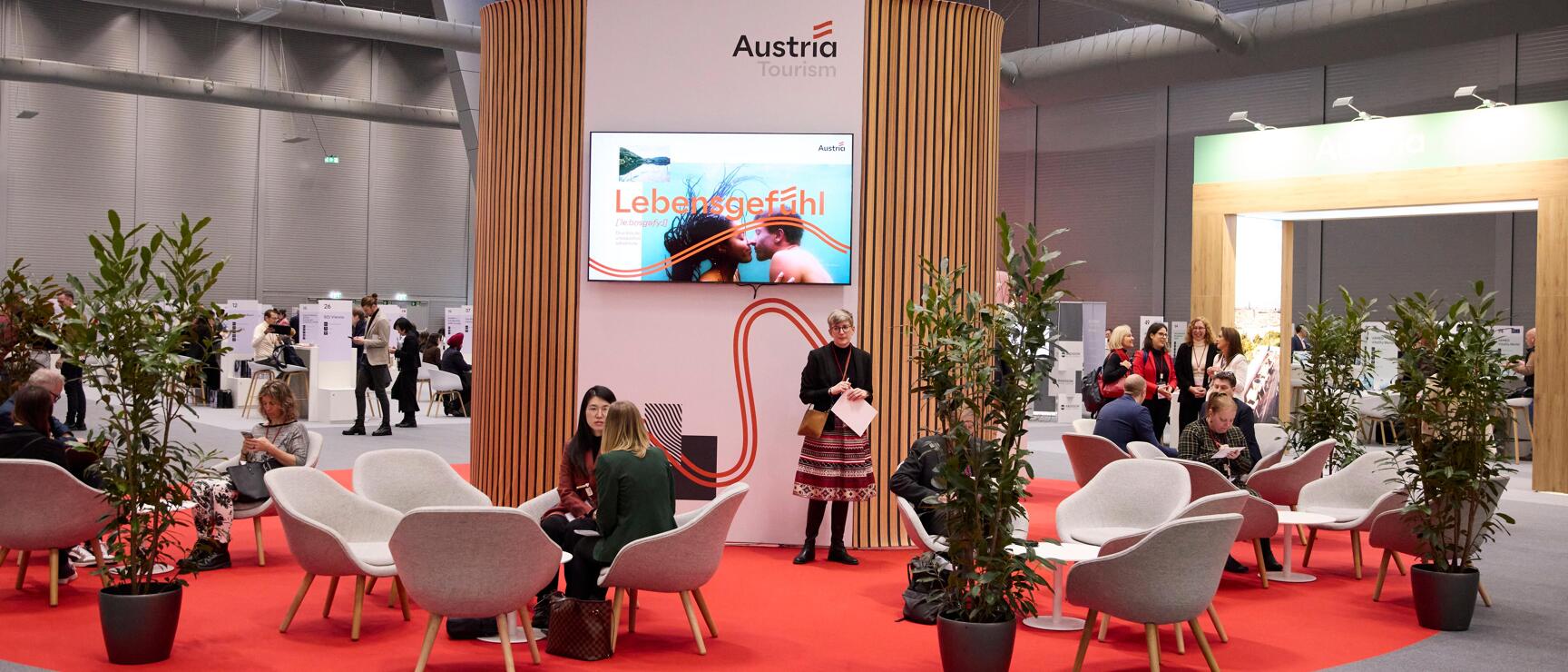 ATB trade fair booth with “Austria Tourism” logo, red carpet, lounge chairs, plants, and visitors in conversation.
