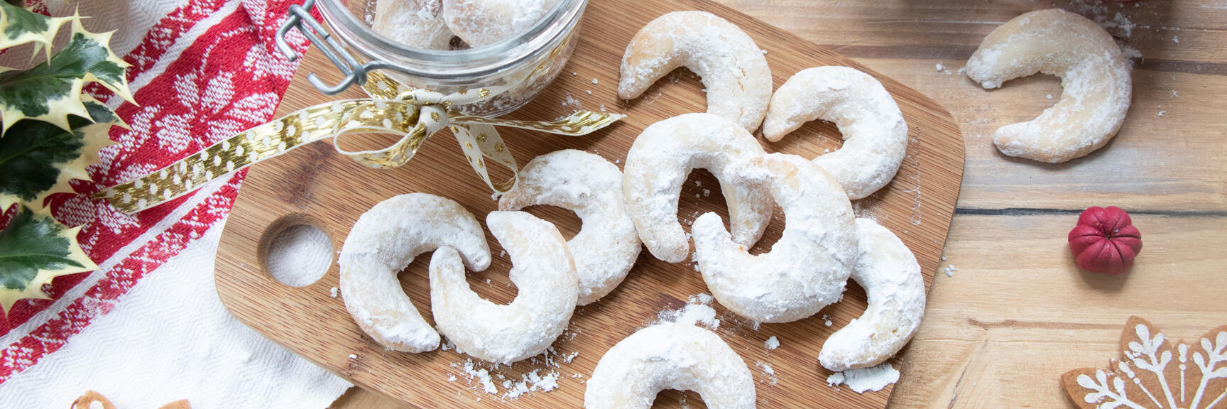 Vanilla crescents with icing sugar on wooden board, jam jar, candles and holly in background.