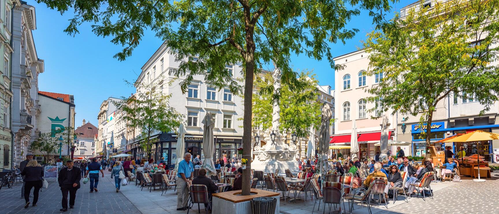 Busy pedestrian zone with street café seating, trees and Austro-Hungarian architecture in sunny weather in St. Pölten.