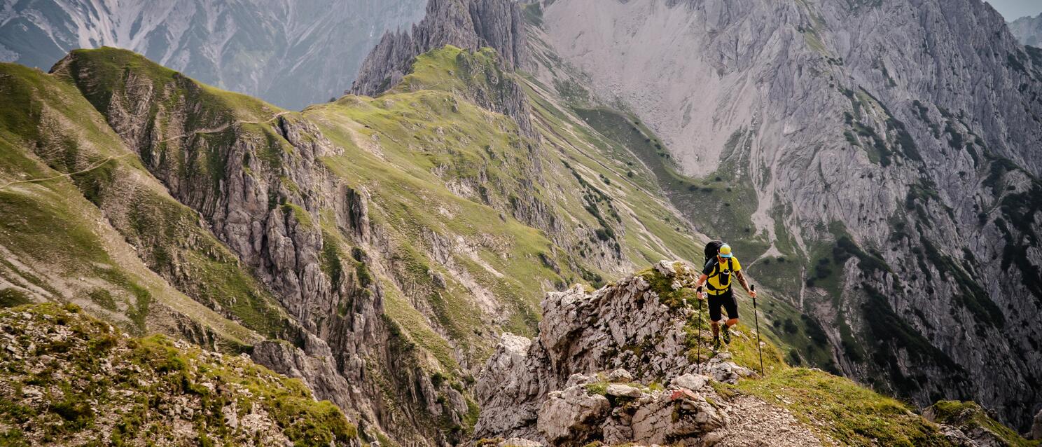 Hiker with yellow backpack and trekking poles on rocky ridge with alpine peaks in background