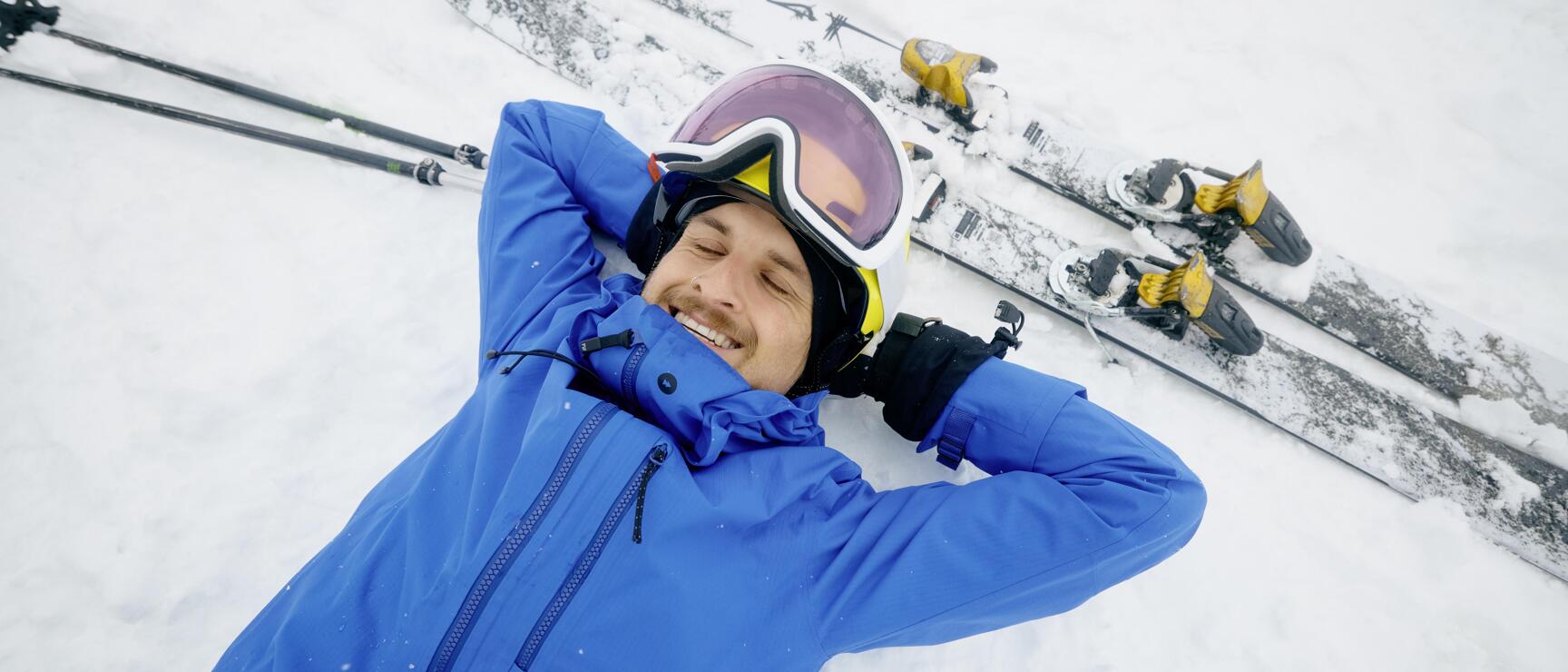 Mann in blauer Skijacke mit Helm und Skibrille liegt lächelnd im Schnee, Skier und Stöcke neben ihm.