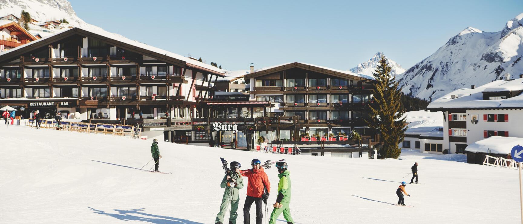 Three skiers on snowy slope in front of Hotel Burg, snow-covered mountains in background, Lech am Arlberg.