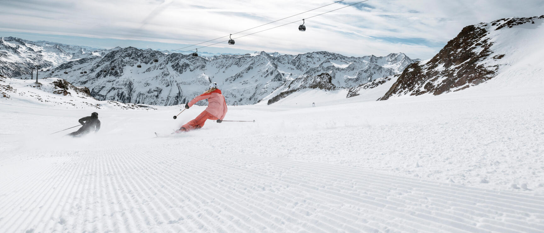 Two skiers on a prepared slope with cable car and snow-covered mountain range in the background