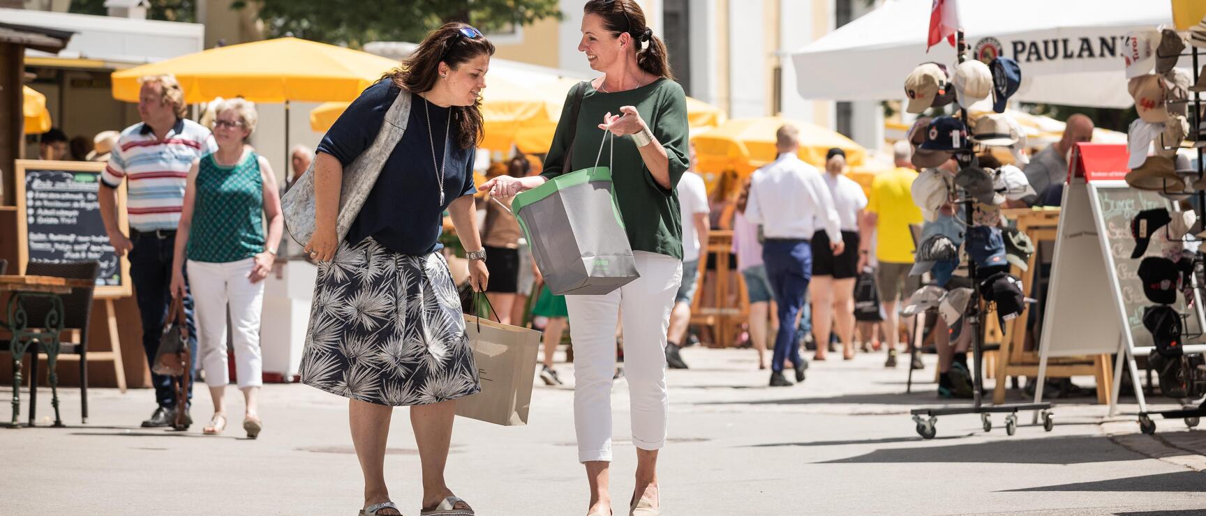 Two women with shopping bags on a busy marketplace, yellow parasols and market stalls in the background.