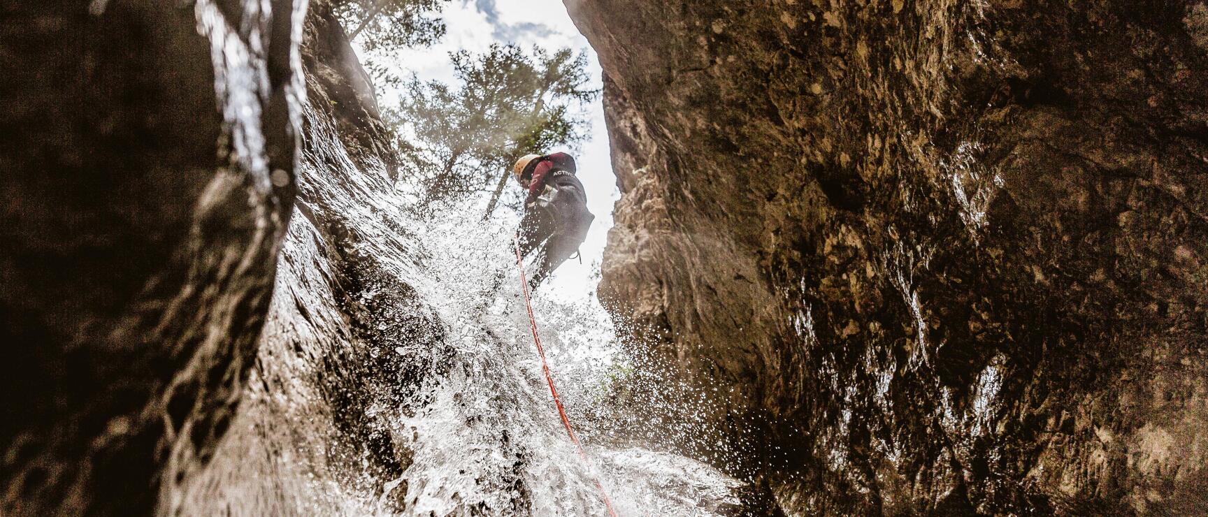 Person wearing helmet abseiling down a waterfall through a narrow rock gorge with red rope visible.