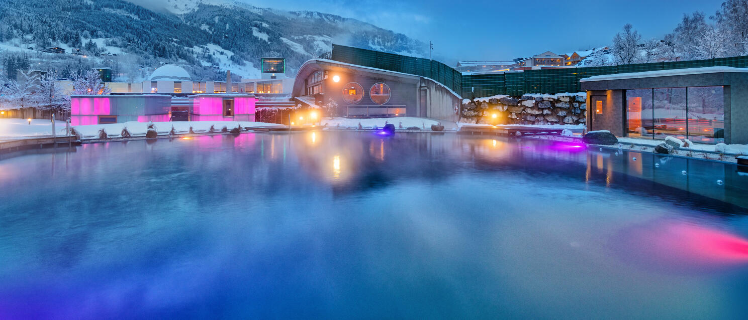 Illuminated outdoor pool of a thermal bath building in winter, snow-covered mountains in the background, dusk.