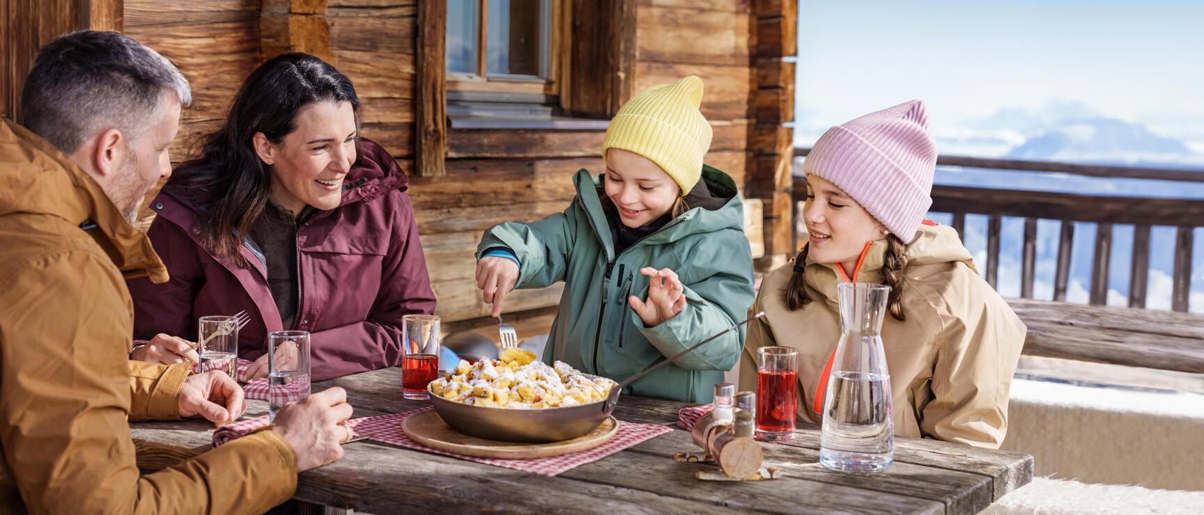 Two adults and two children eating kaiserschmarrn on an alpine hut terrace, snow-covered mountains in the background.