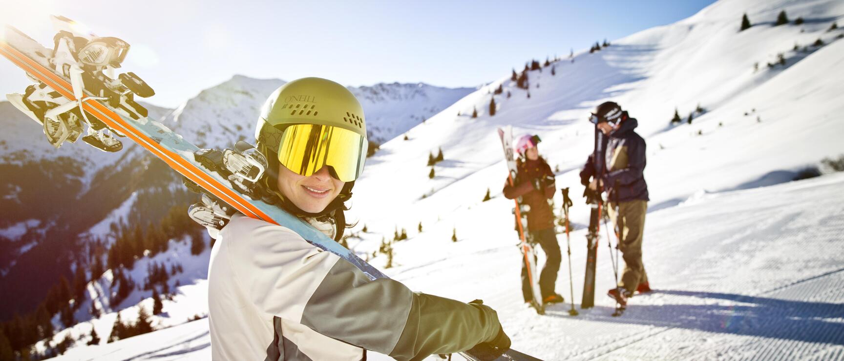 Person with ski helmet and goggles carries skis over shoulder, two other skiers in snowy mountain terrain.