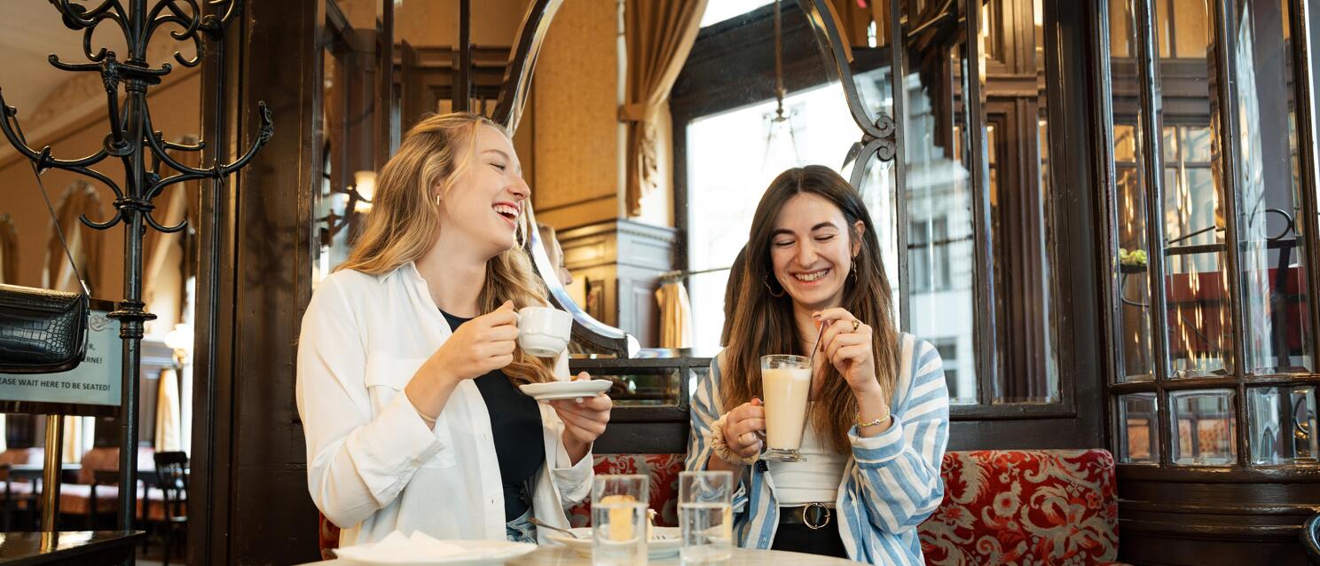 Two laughing women with coffee at a marble table in a Viennese coffee house.
