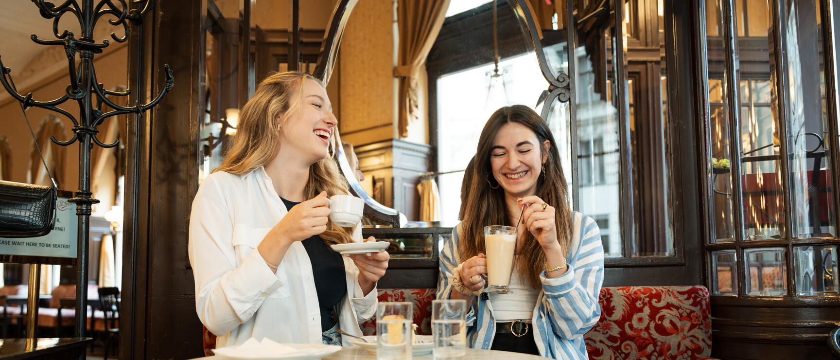Two laughing women with coffee at a marble table in a Viennese coffee house.