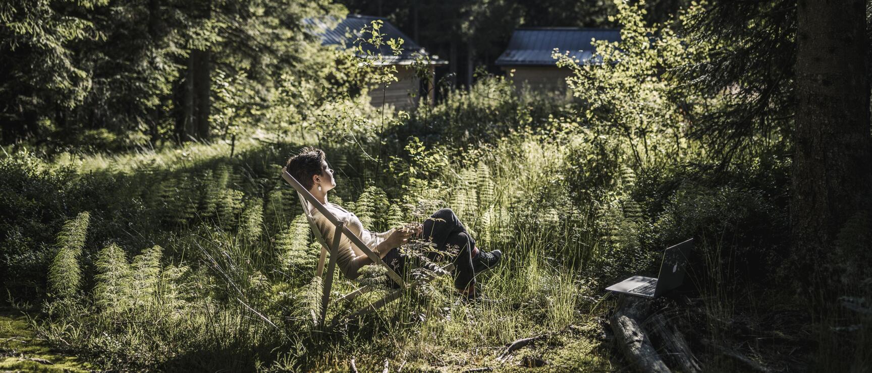 Person in deckchair in coniferous forest with laptop nearby and wooden cabin in background