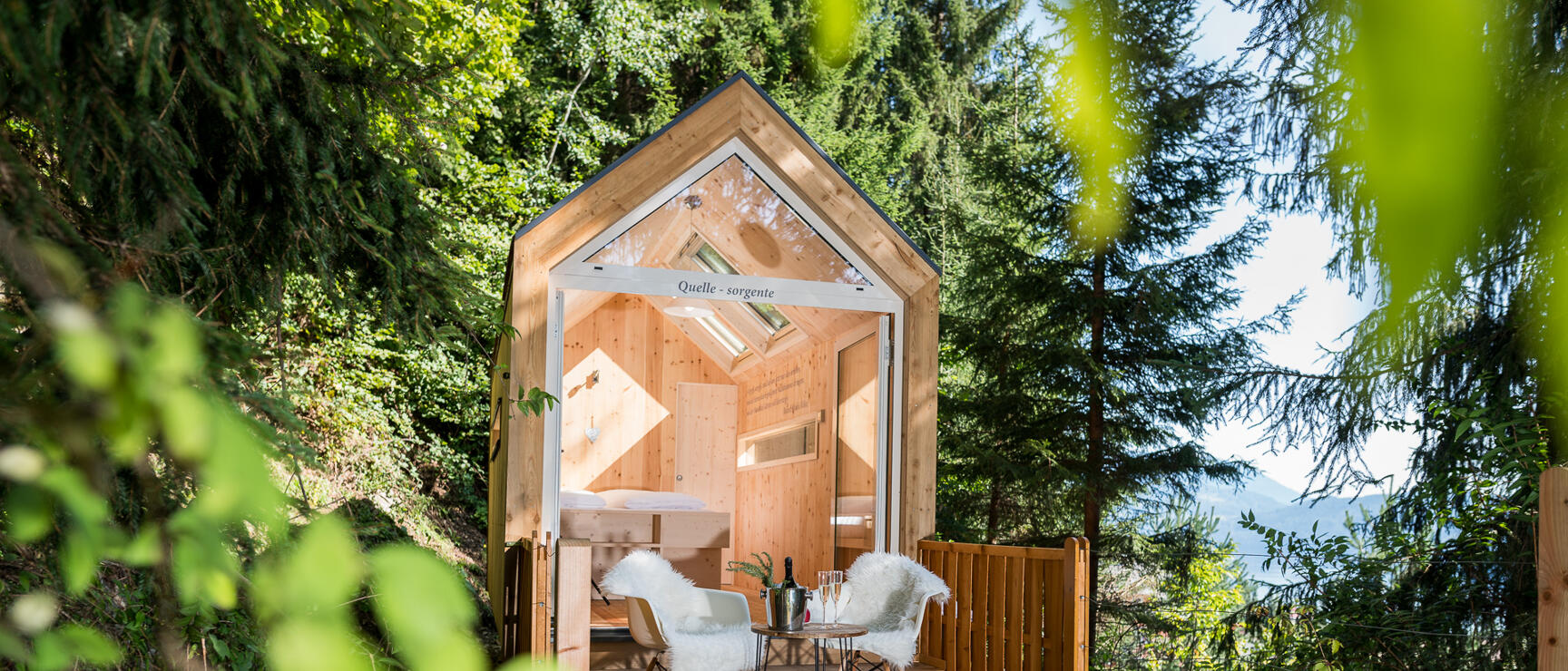 Small wooden cabin with wooden terrace, two chairs and champagne glasses in forest, mountains in background.