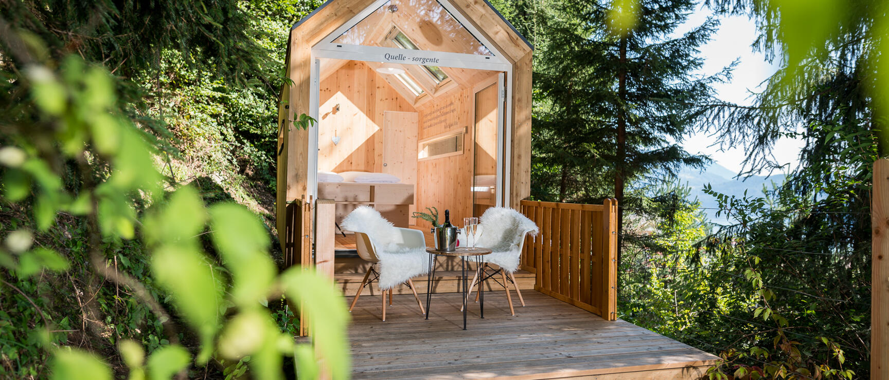 Kleines Holzhäuschen mit Holzterrasse, zwei Stühlen und Sektgläsern im Wald, Berge im Hintergrund.