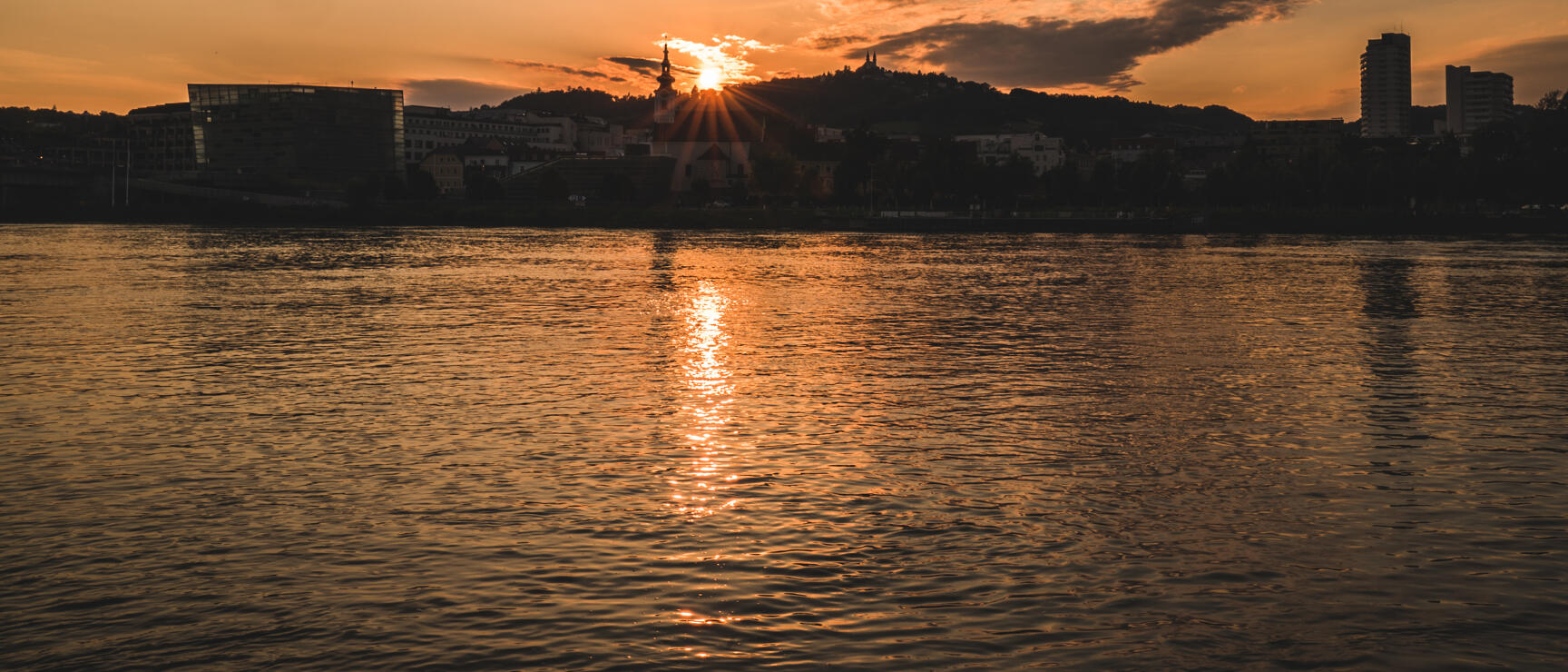 Sunset over Linz with city skyline, church tower and Pöstlingberg reflected in Danube water.