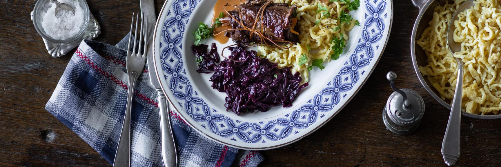 Beef roulade with gravy, red cabbage and egg noodles on blue and white plate with side dishes on wooden table.