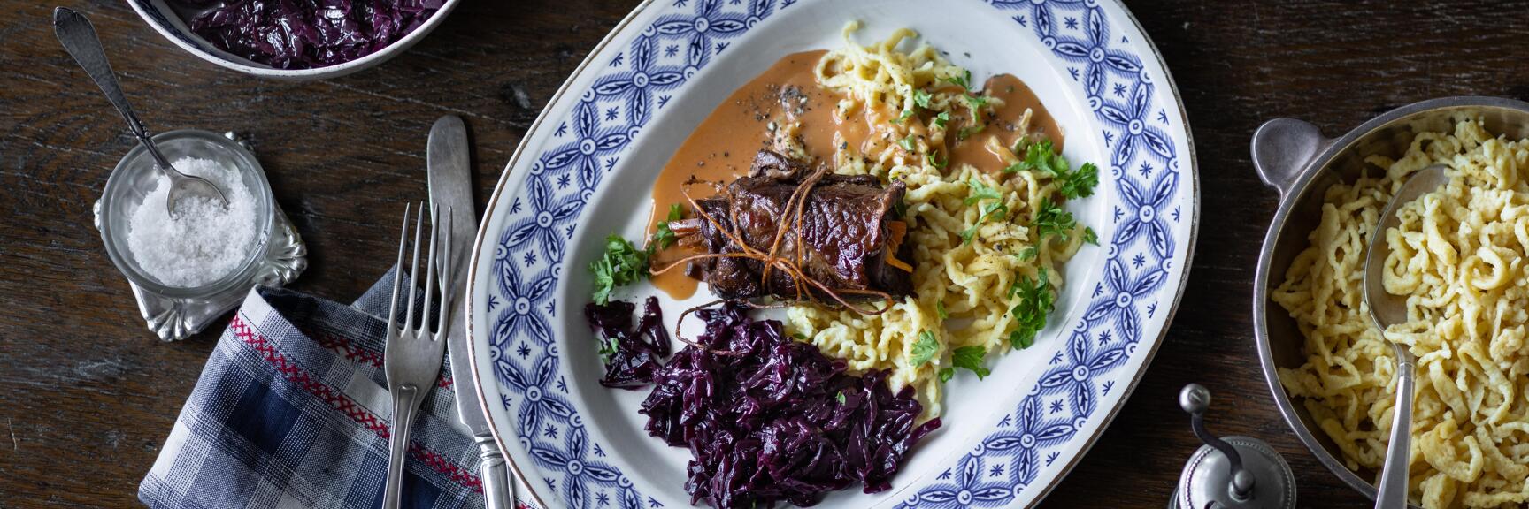 Beef roulade with gravy, red cabbage and egg noodles on blue and white plate with side dishes on wooden table.