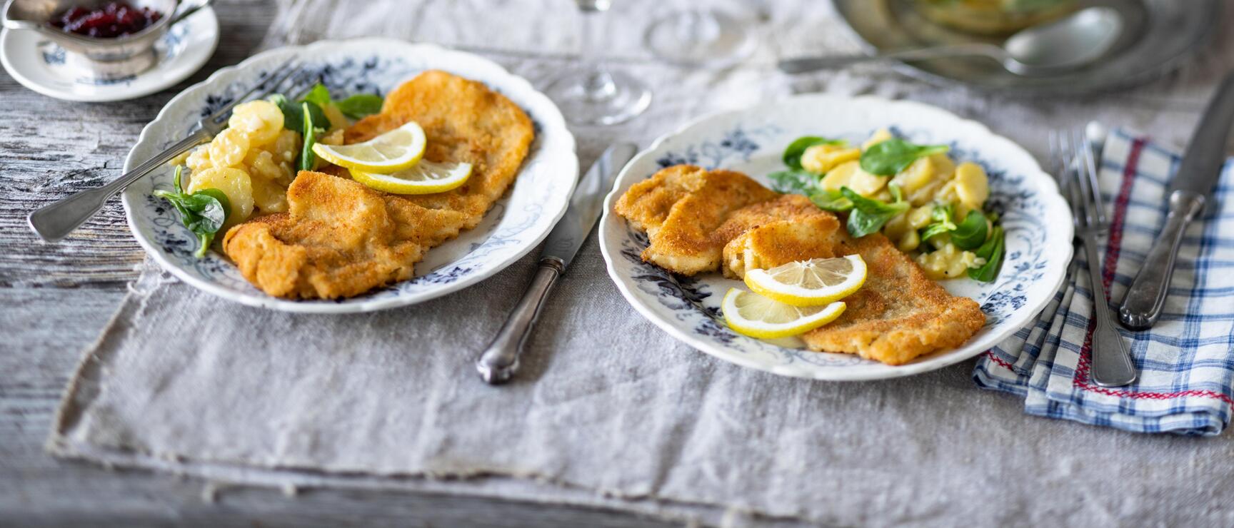 Two plates of Wiener schnitzel with lemon slices and potato salad, two white wine glasses in the background.