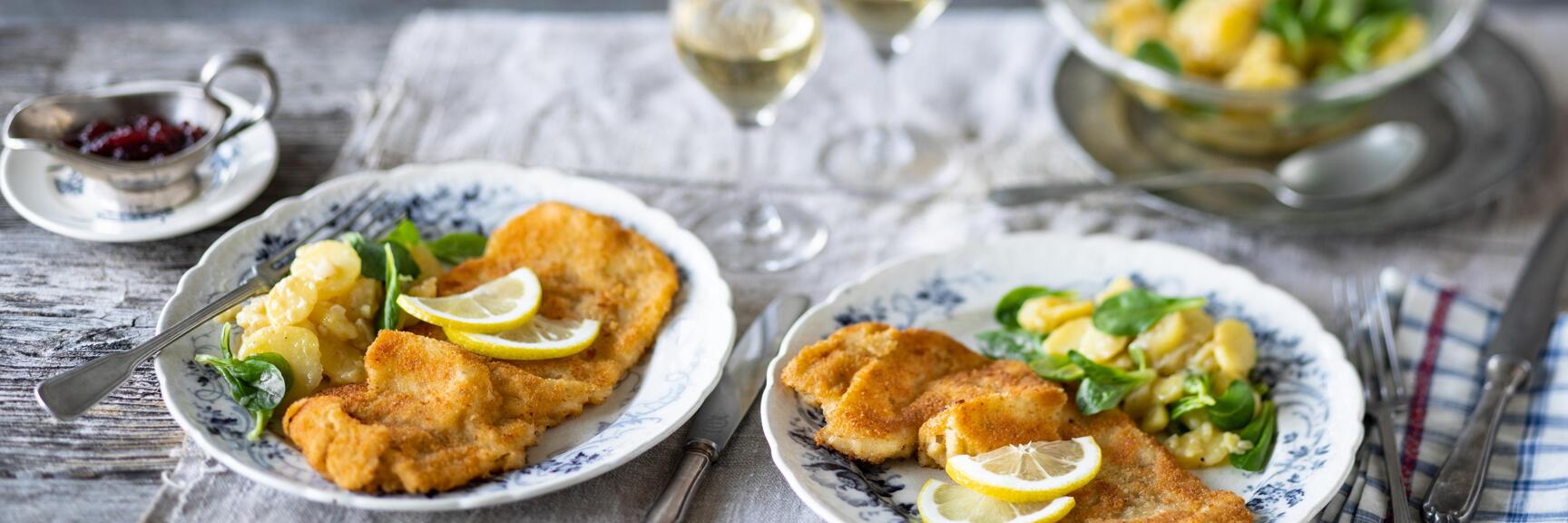 Two plates of Wiener schnitzel with lemon slices and potato salad, two white wine glasses in the background.