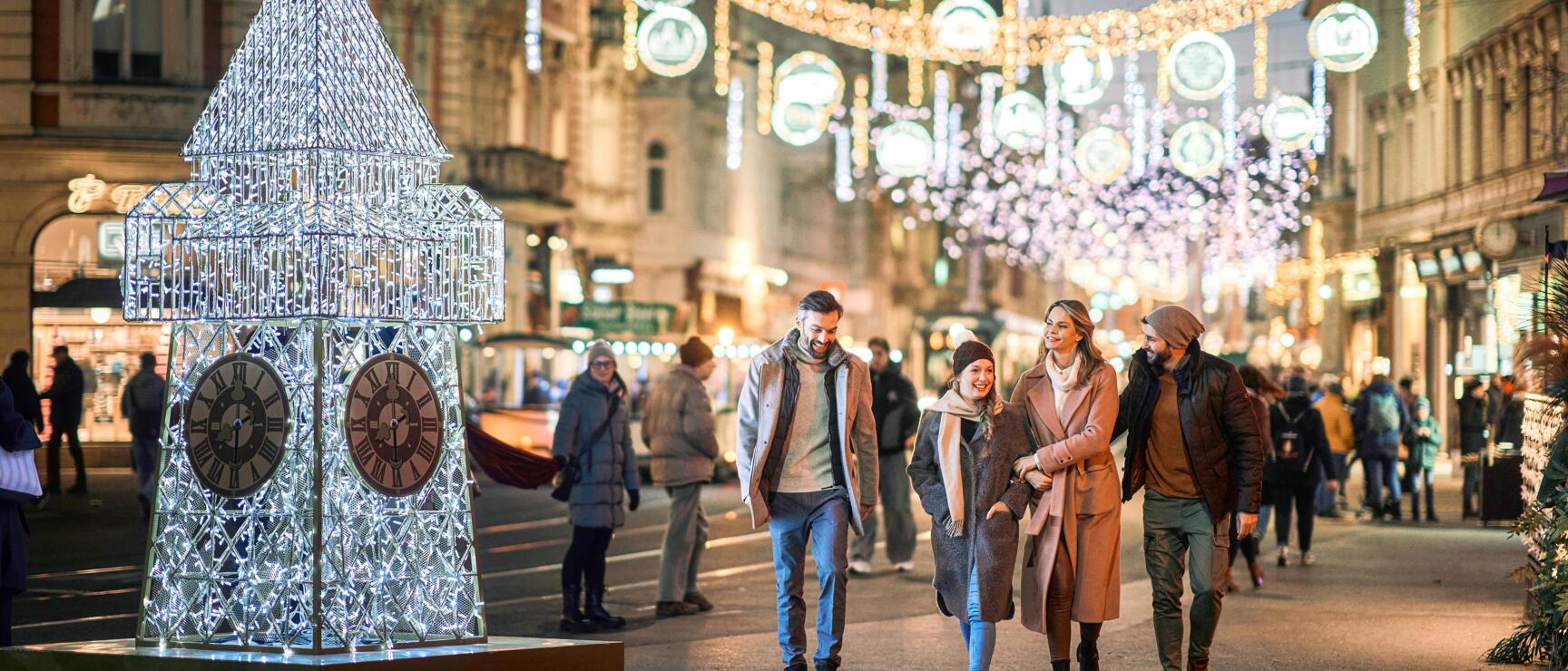 Eine Familie spaziert neben der Glitzernden Uhrturm- Figur im festlich geschmückten Graz