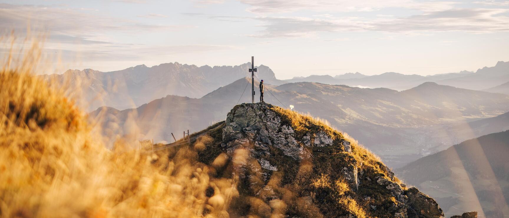 Person neben Gipfelkreuz auf felsigem Berggipfel, weites Alpenpanorama im Hintergrund, goldenes Gras im Vordergrund.