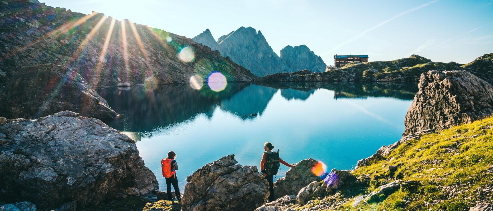 Two hikers with backpacks at a mountain lake, rocky peaks and alpine hut in the background, blue sky.