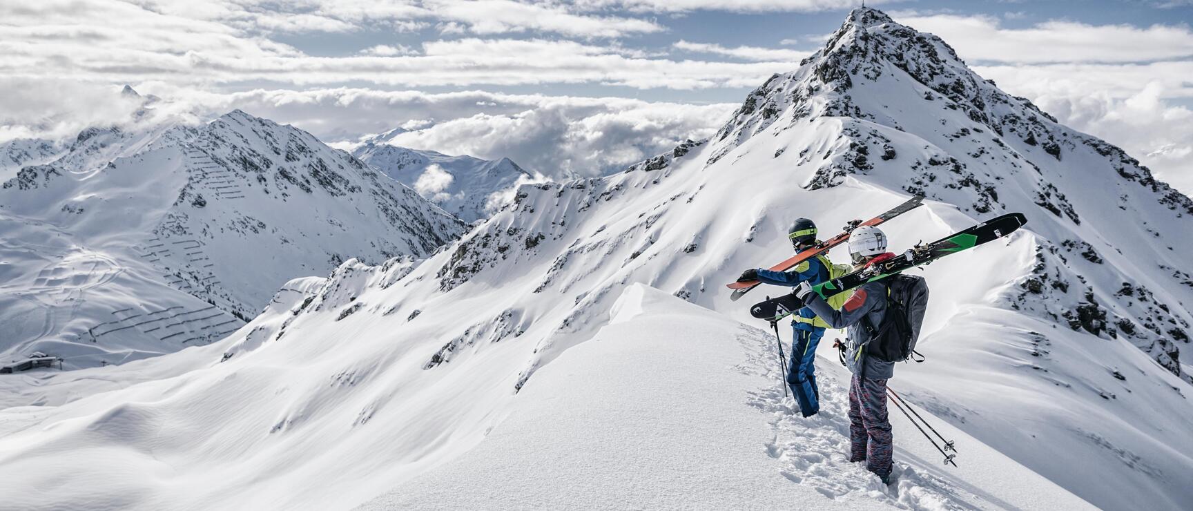 Two people carrying skis on their shoulders walking along a snowy mountain ridge, summit cross in the background.