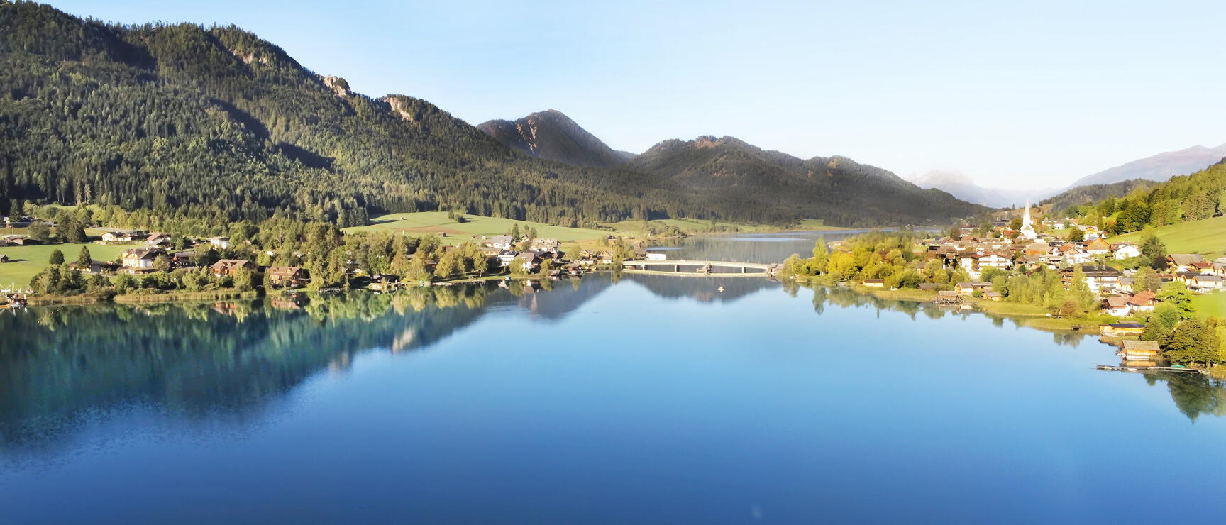 Calm mountain lake with reflection, village with church on shore, forested mountains in background.