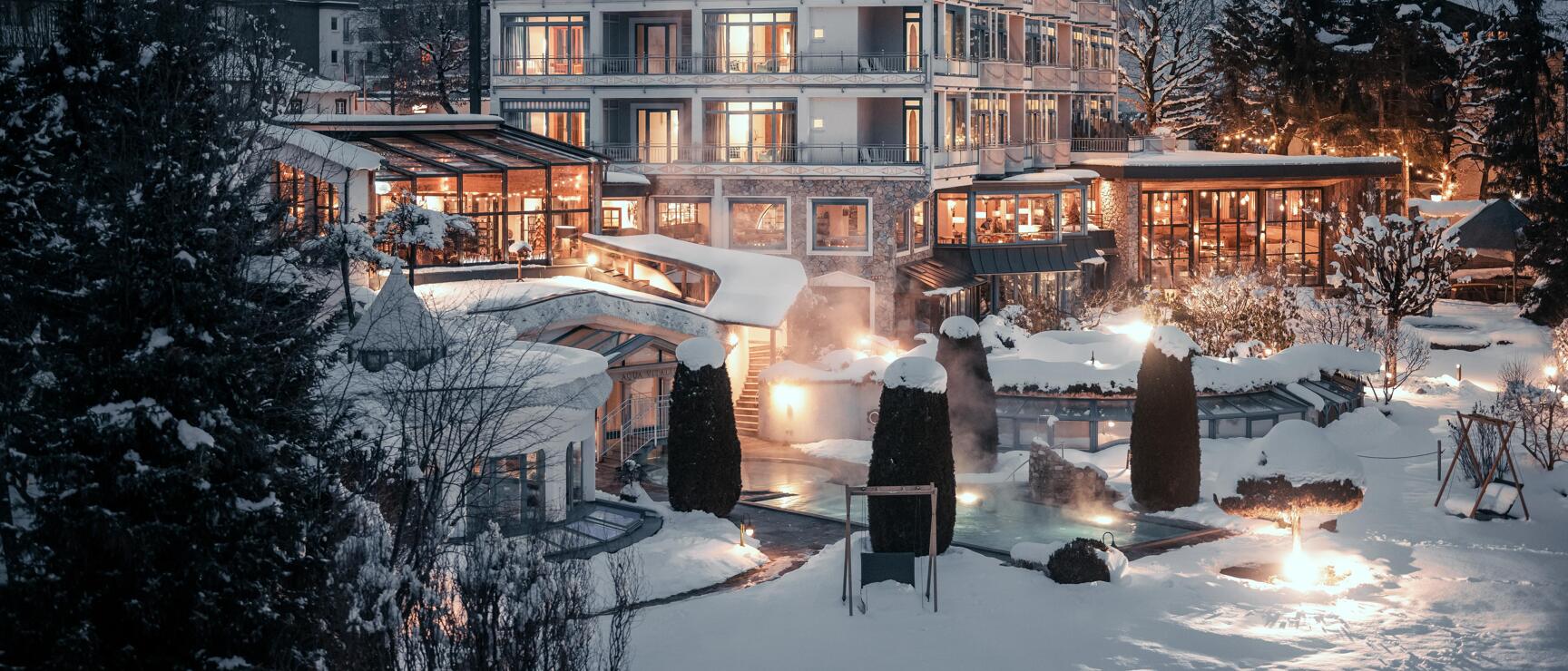 Illuminated hotel building in winter at dusk, outdoor pool with steam, snow-covered mountain slope in the background.