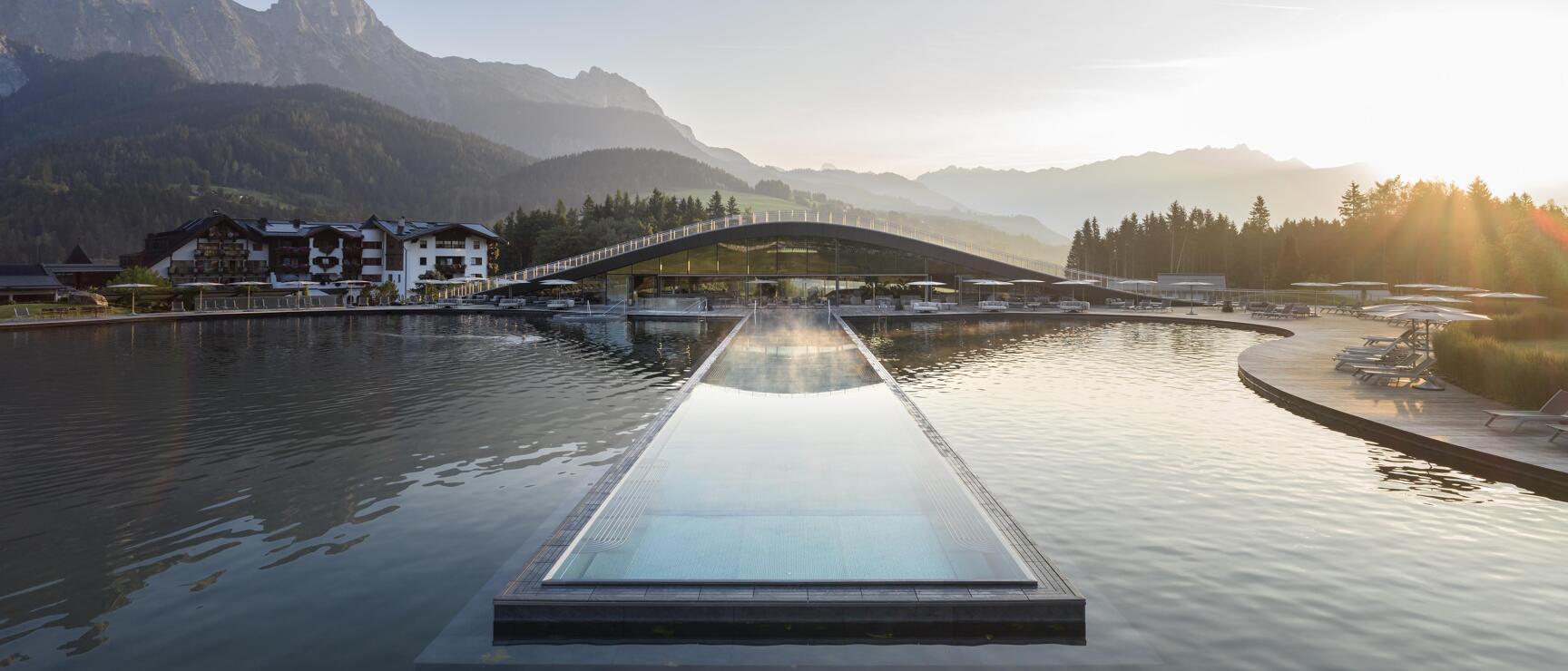 Infinity pool on natural lake, modern wellness building and mountain panorama in morning light.