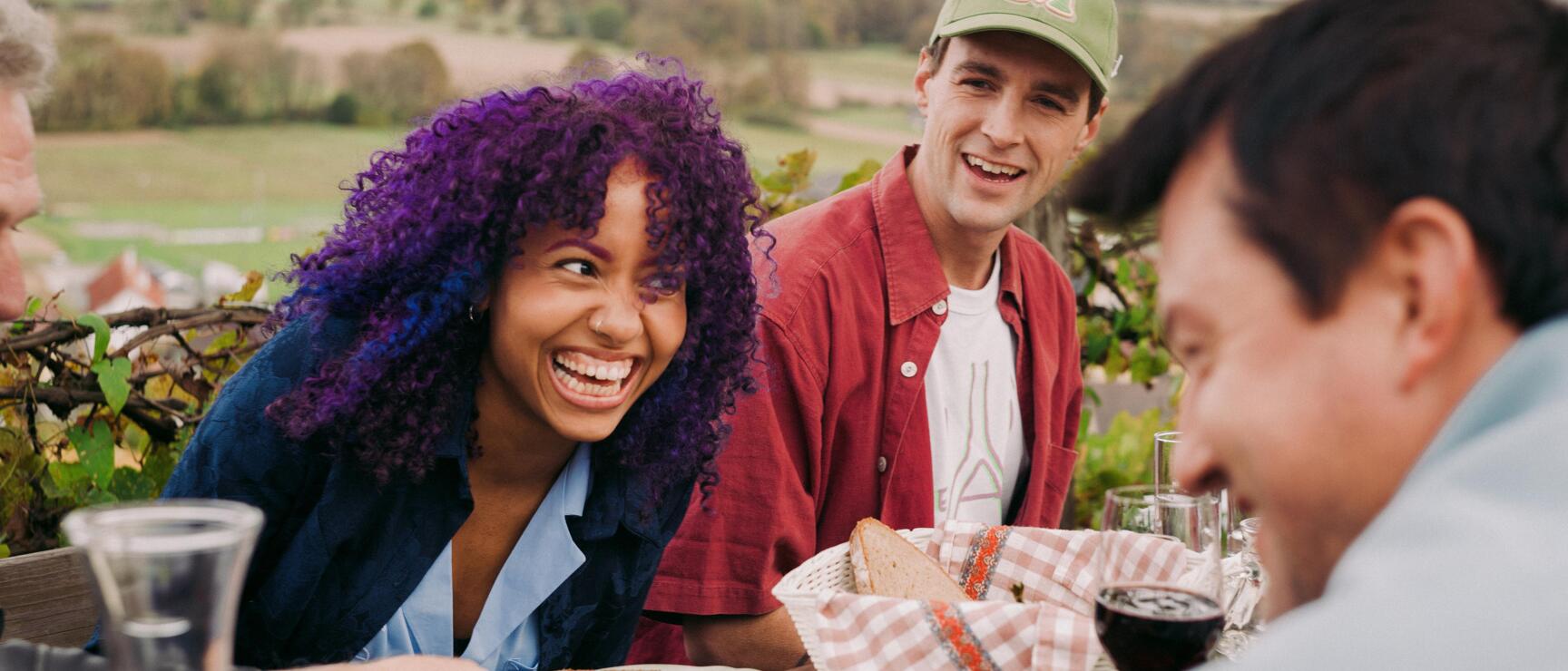 Three people laughing at a set table outdoors, bread and red wine, hilly landscape in the background.