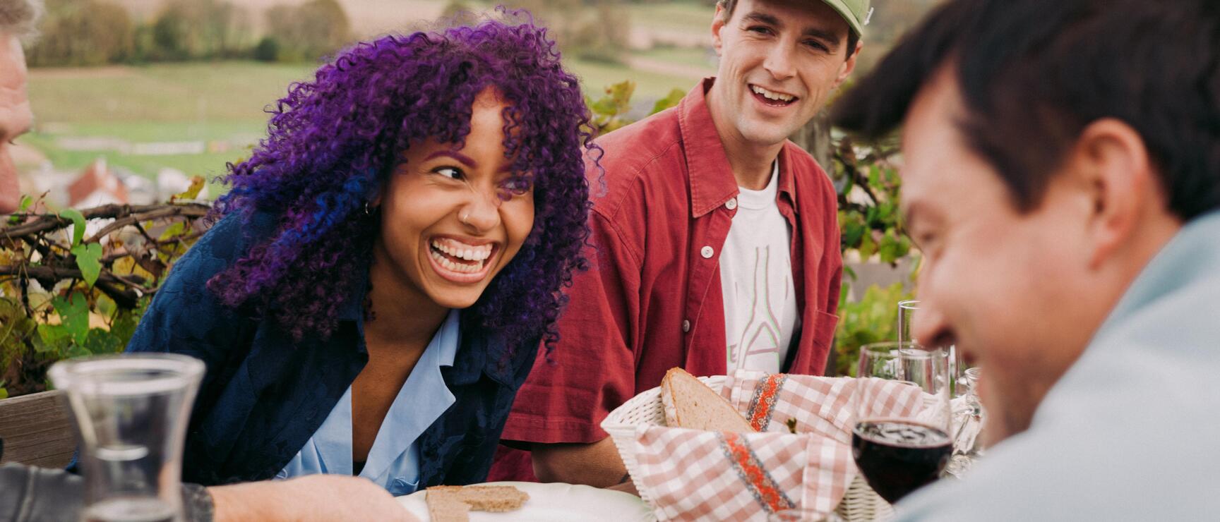 Drei Personen lachen an einem gedeckten Tisch im Freien, Brot und Rotwein, Hügellandschaft im Hintergrund.