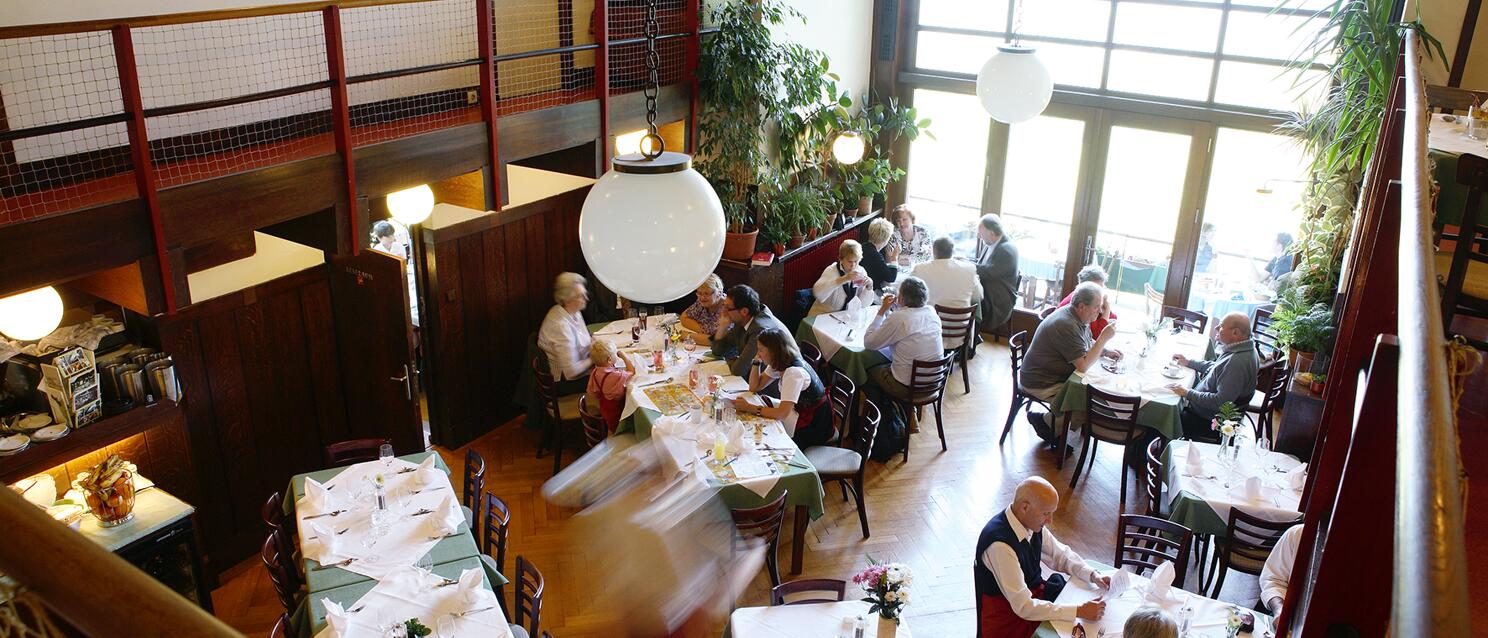 Overhead view of a Viennese pub with laid tables, guests, waiter, and gallery in the background.