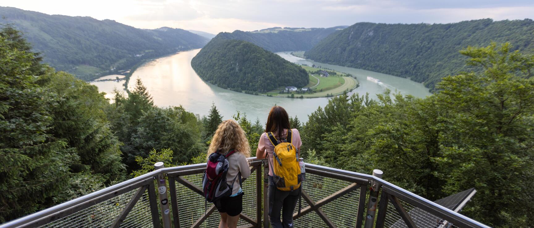 Two women with backpacks on a metal observation platform overlooking a Danube bend with forested hills.