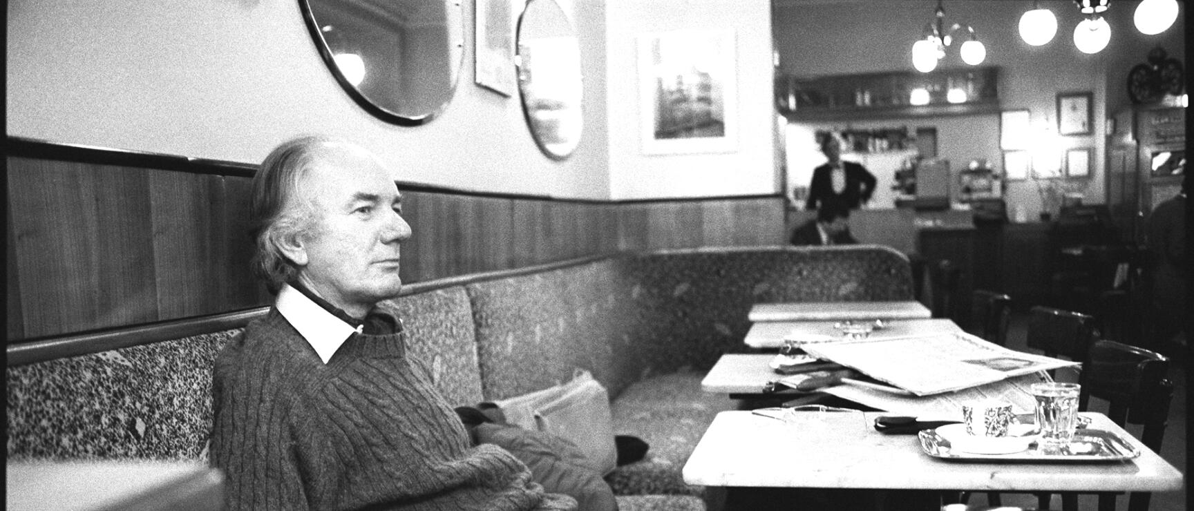 Man in knit sweater sitting on an upholstered bench in a Viennese coffee house, newspapers and glasses on a marble table.