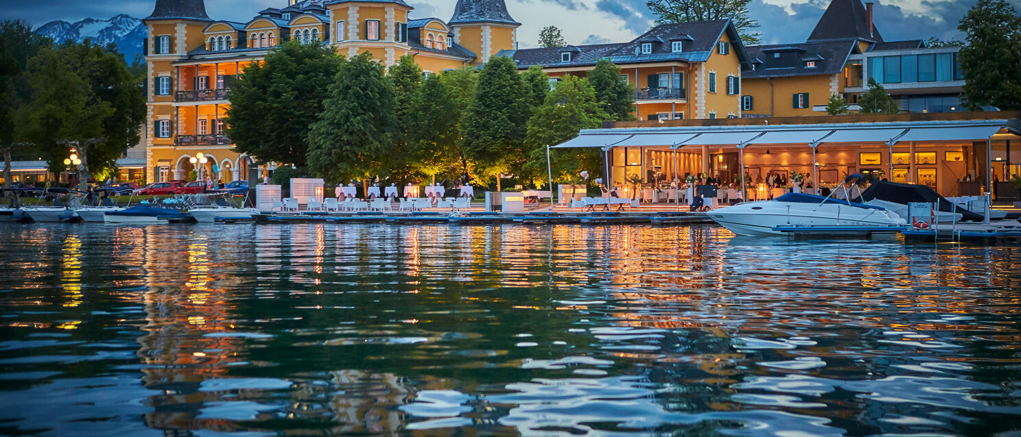 Velden Castle on Lake Wörth at dusk, illuminated lakeside terrace, boats in foreground, mountains in background.