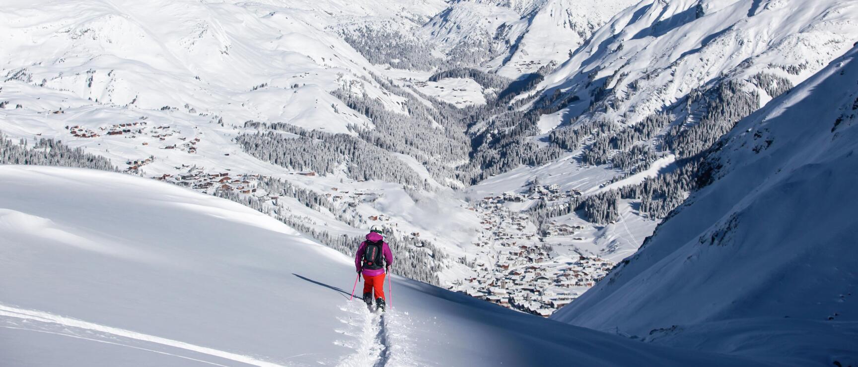Ski tourer in pink jacket on snowy slope with view of Lech am Arlberg and mountain panorama.