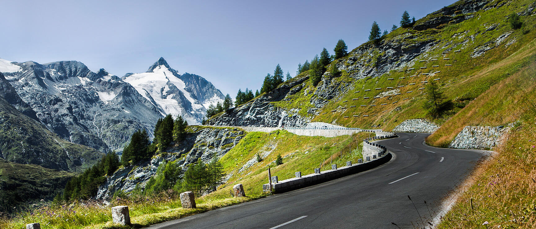 Winding high alpine road with stone bollards, green mountainside, snow-capped peaks in background.