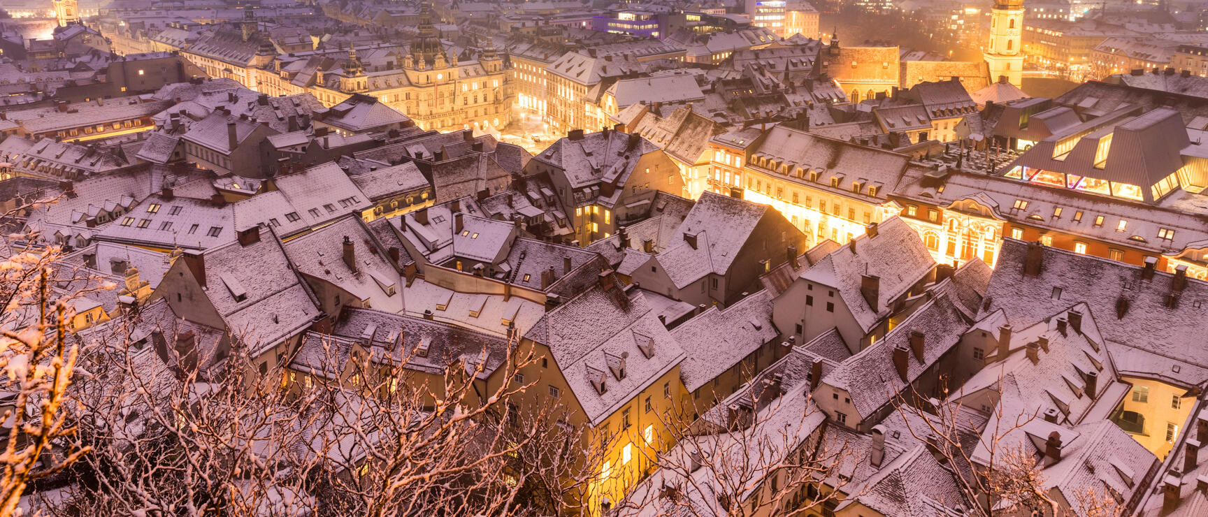Winter view over snow-covered roofs of Graz old town at night, illuminated town hall.