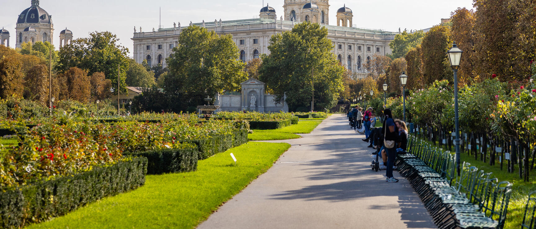 View from Volksgarten to Kunsthistorisches Museum Vienna, park path with roses and benches, people sitting