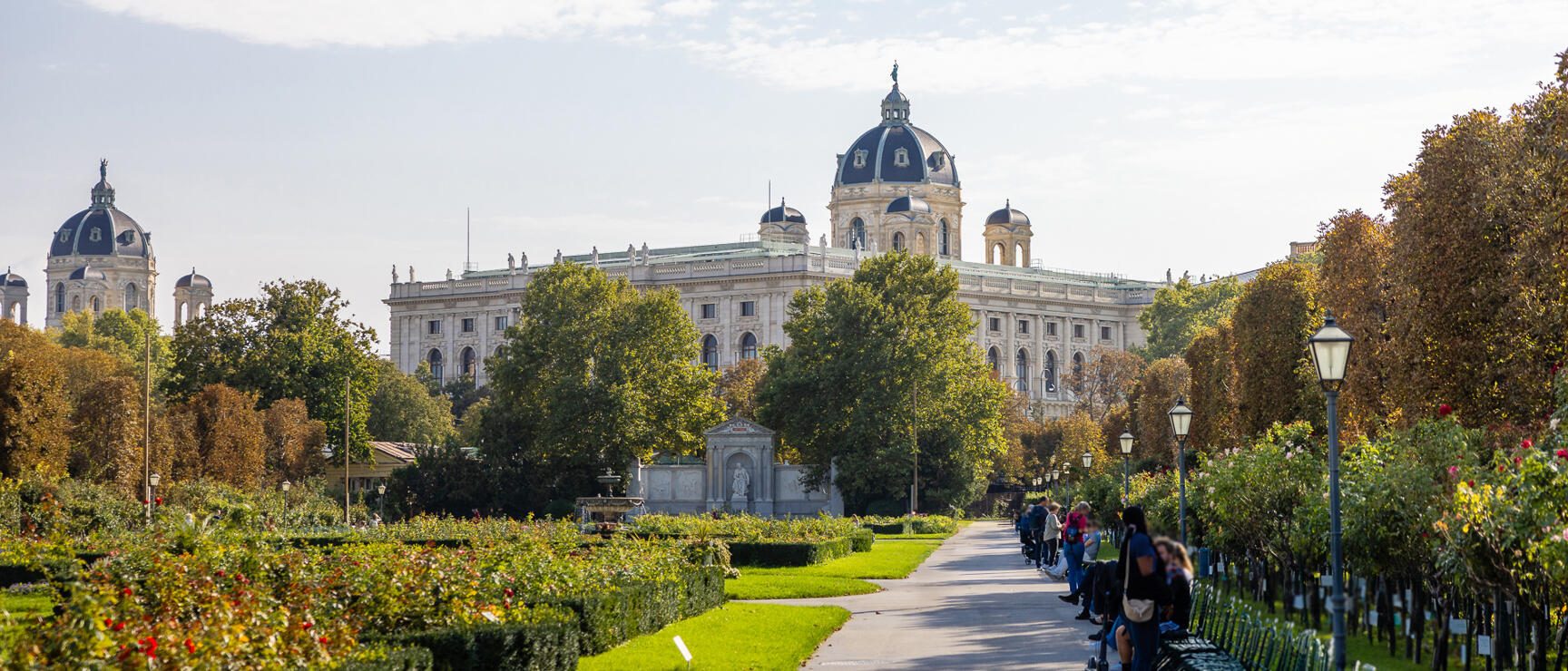 Blick vom Volksgarten auf das Kunsthistorische Museum Wien, Parkweg mit Rosen und Bänken, Menschen sitzen.