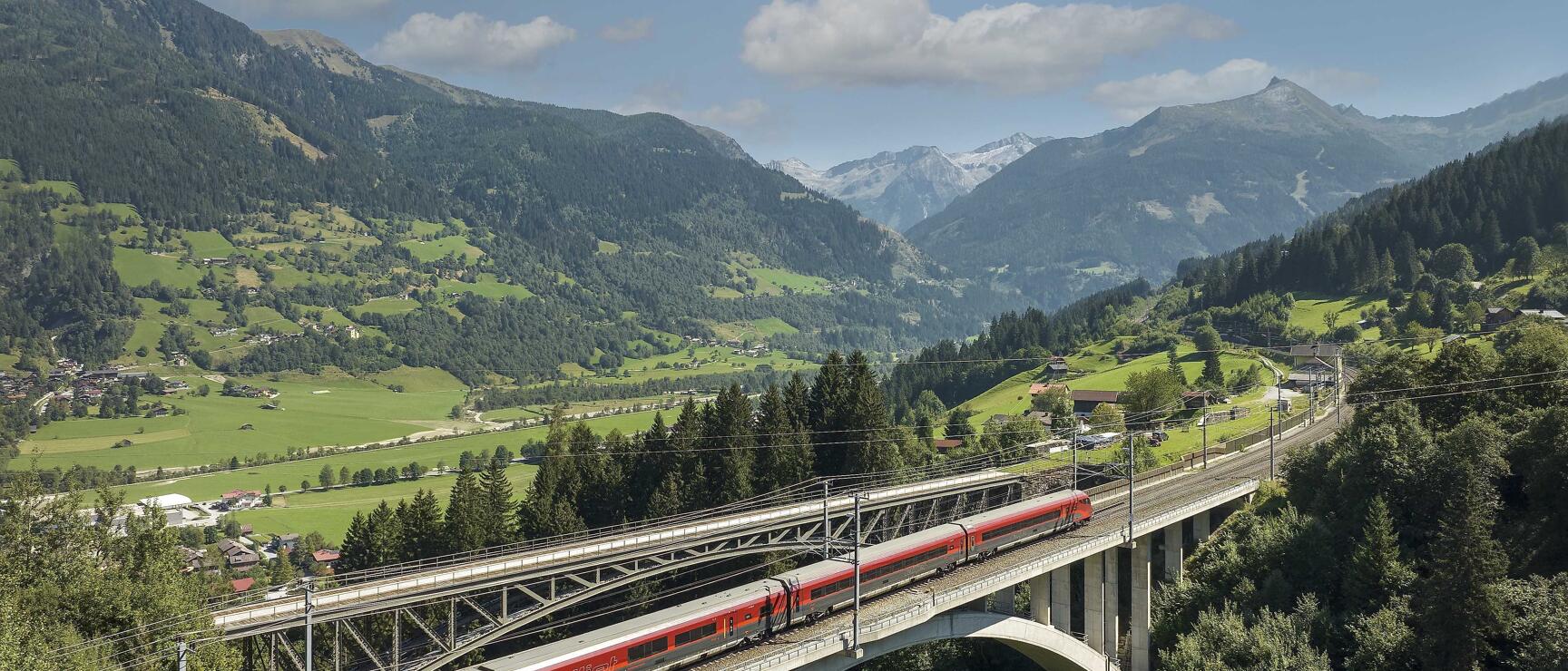 Roter ÖBB-Zug auf Betonbrücke über bewaldeter Schlucht, grünes Alpental mit Bergen im Hintergrund.